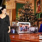Queen Elizabeth is seen sitting behind a table while 4 framed pictures of royal family members are placed on the table.